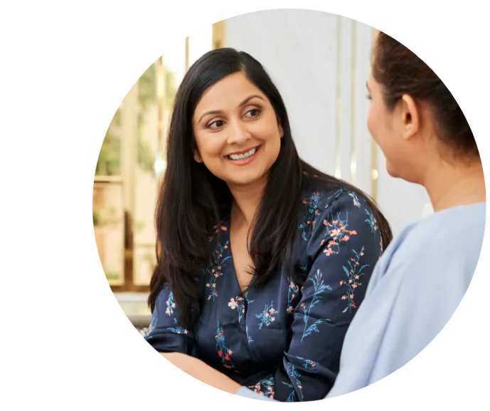 Two women sitting and conversing warmly indoors. The woman on the left, wearing a floral-patterned top, is smiling, creating a friendly, relaxed atmosphere.